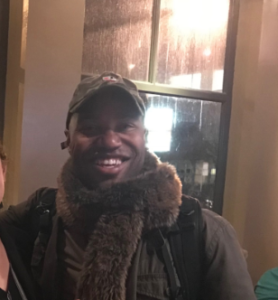 jovan johnson in a hat, scarf, and jacket that are all colored in green and brown earth tones